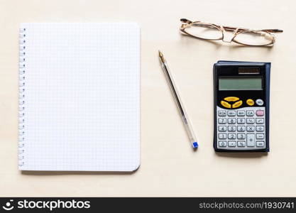 top view of blank sheet of squared paper of spiral notebook, pen, eyeglasses and financial calculator on light brown wooden board