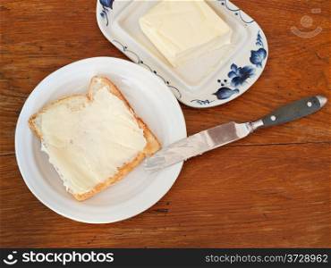 top view of bread and butter sandwich on white plate, knife, dairy butter, butterdish on wooden table