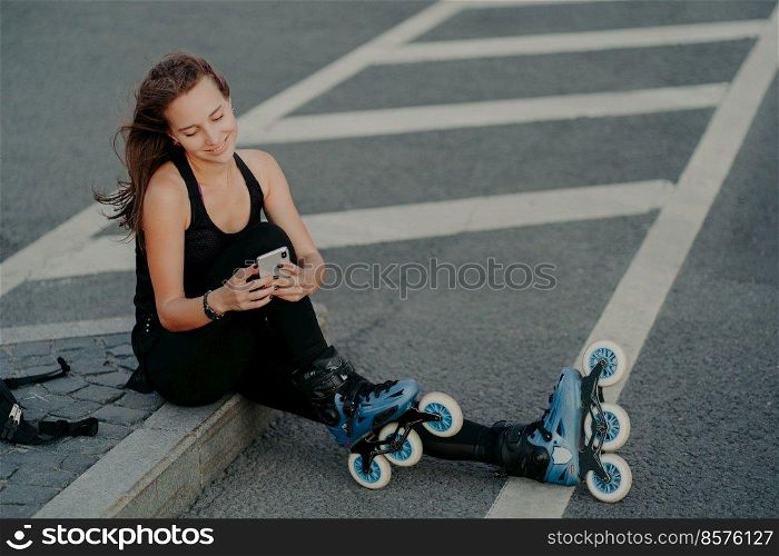 Top view of brunette active young woman uses smartphone for online communication poses on asphalt dressed in black active wear enjoys favorite hobby and free time. Extreme sport and rest concept