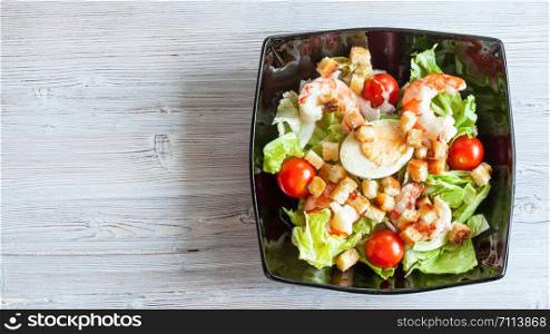 top view of Cesar salad with prawns in black bowl on gray wooden table with panoramic copyspace