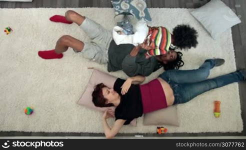 Top view of cheerful interracial family having fun while lying on the floor at home. Playful african dad raising his mixed race adorable son up into the air over his head while caucasian mom relaxing and smiling. Overhead. Slow motion.