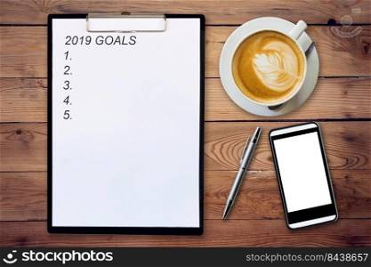 Top view of clipboard written Goals 2019 on wooden background with coffee cup, pen and mobile phone.