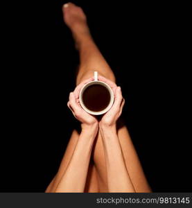 Top view of crop unrecognizable barefooted female sitting in black studio with cup of hot coffee. Crop anonymous woman with coffee cup in black studio