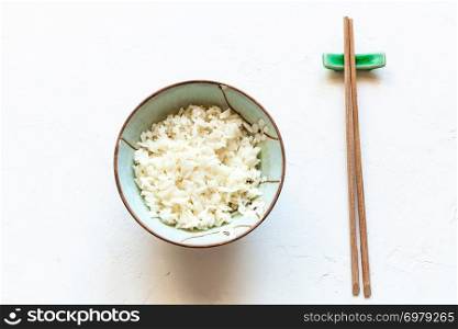 top view of cup with boiled rice and wooden chopsticks on rest on white concrete board