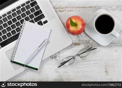 Top view of desktop with partial laptop, apple, coffee, reading glasses, and pen.