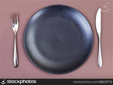 top view of empty black plate with fork and knife on brown background