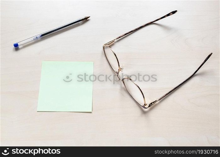 top view of eyeglasses, plastic blue pen on blank green sheet of note paper on light brown wooden board