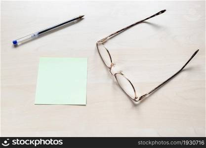 top view of eyeglasses, plastic blue pen on blank green sheet of note paper on light brown wooden board