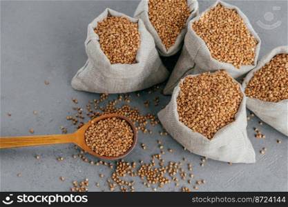 Top view of five small sacks filled with raw buckwheat, wooden spoon near with uncooked cereals, dark background. Healthy food concept