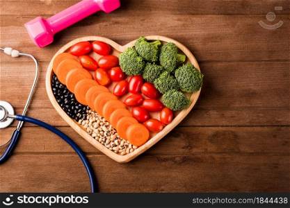 Top view of fresh organic fruits and vegetables in heart plate wood (carrot, Broccoli, tomato), doctor stethoscope and sport dumbbell on wooden table, Healthy lifestyle diet food concept