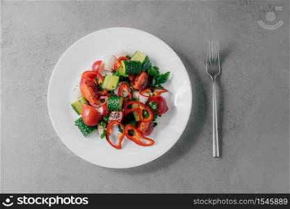 Top view of fresh vegetable salad prepared of red pepper, radish, tomatoes, cucumbers and parsley in white bowl, fork near. Vegetarian dish concept. Healthy nutritious spring salad