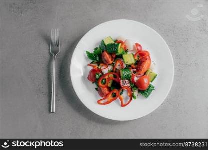 Top view of fresh vegetable salad prepared of red pepper, radish, tomatoes, cucumbers and parsley in white bowl, fork near. Vegetarian dish concept. Healthy nutritious spring salad