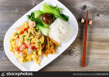 Top view of freshly cooked egg and shrimp dish with bok choy, soy sauce egg, cucumber, rice and chopsticks in holder on rustic wood.