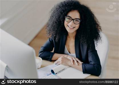 Top view of glad African American businesswoman smiles pleasantly, notes information, makes notes, wears transparent glasses, formal clothes, computer monitor in front, sits at desktop alone