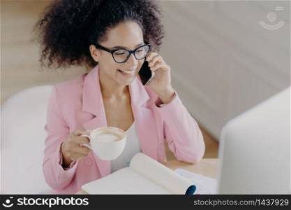 Top view of glad dark skinned woman with curly dark hair, has phone conversation, holds mug of drink, smiles pleasantly while looks at screen of computer, wears elegant attire, busy working at project