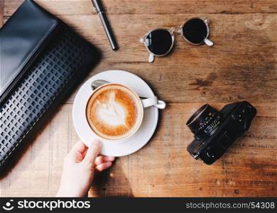 Top view of hand holding hot coffee on wooden table with woman bag with camera and sunglasses