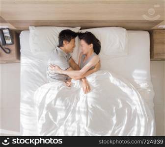 Top view of happy Asian couple smiling, and sleeping together on bed in love and sex concept in a modern bedroom with white blanket in the morning.