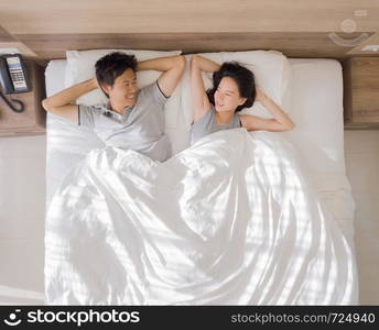 Top view of happy Asian couple smiling, and sleeping together on bed in love and sex concept in a modern bedroom with white blanket in the morning.