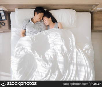 Top view of happy Asian couple smiling, and sleeping together on bed in love and sex concept in a modern bedroom with white blanket in the morning.