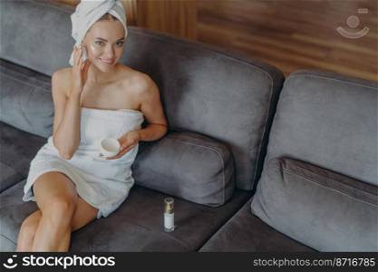 Top view of happy Caucasian woman satisfied with healthy smooth perfect skin, applies face cream, wrapped in bath towel after taking shower, poses on couch at home. Freshment, wellness concept