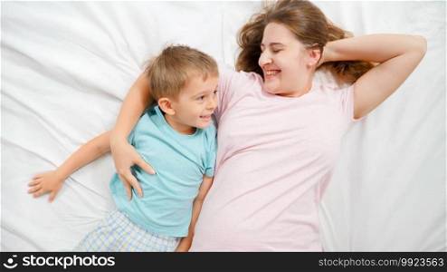 Top view of happy cheerful young mother with little toddler son in pajamas lying on bed and smiling. COncept of parenting and family happiness.. Top view of happy cheerful young mother with little toddler son in pajamas lying on bed and smiling. COncept of parenting and family happiness