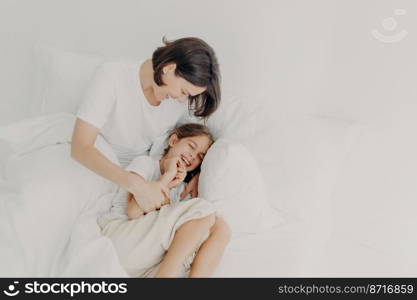 Top view of happy funny girl in pyjamas and her caring mom pose on bed, play together in bed, enjoy softness and comfort, express positive emotions. Family, morning time and relaxation concept