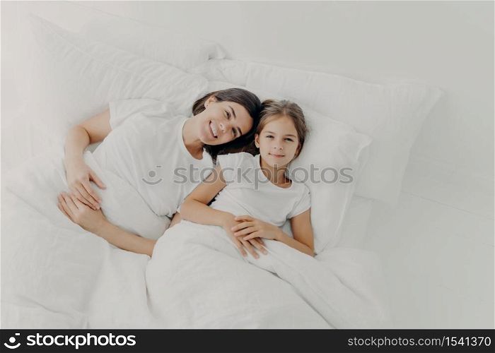 Top view of happy mother and daughter awake in good mood, feel relaxed, enjoy comfort in bed, rejoice good morning, dressed in pyjamas, lie under white blanket on soft pillows. Bedding time.