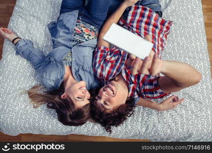 Top view of happy young couple in love taking a selfie with smartphone lying over a bed. Leisure time at home concept.. Couple in love taking selfie with smartphone on bed
