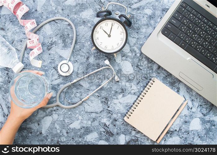 Top view of healthy woman desk with stethoscope, woman&rsquo;s hand holding glass of water and blank notebook with laptop computer. Top view with copy space, flat lay.
