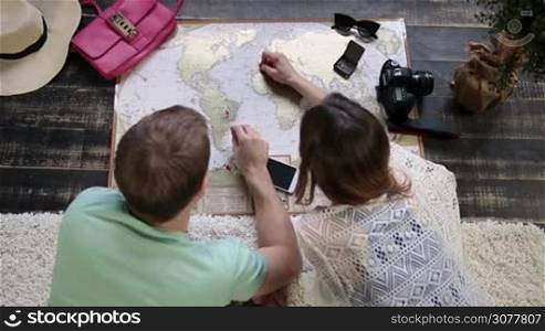 Top view of hipster couple lying on floor at home and planning vacation trip using scrath travel map of the world. Rear view. Young beautiful woman erasing african country with coin and pointing travel destination with pushpins on scratch travel map.