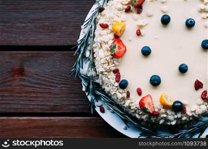 Top view of homemade cake, white chocolate icing decorated fresh berries, goji, citrus and rosemary, brown wooden table background. Image for menu or confectionery catalog