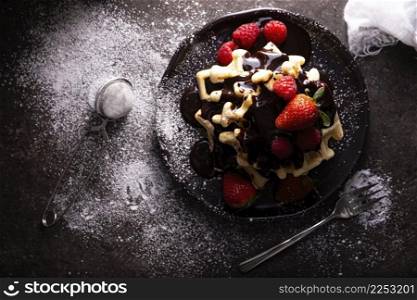 Top view of homemade waffles with chocolate, icing sugar, strawberries and raspberries on dark dish. Messy look flat lay.