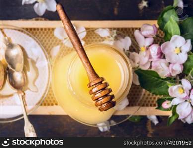 Top view of honey jar with wooden dipper, spoon and spring blossom on honeycomb and rustic wooden background