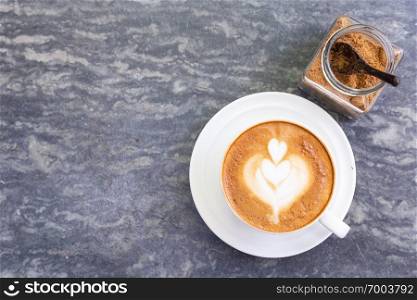 Top view of hot coffee with heart pattern in white cup and brown sugar on stone table background.