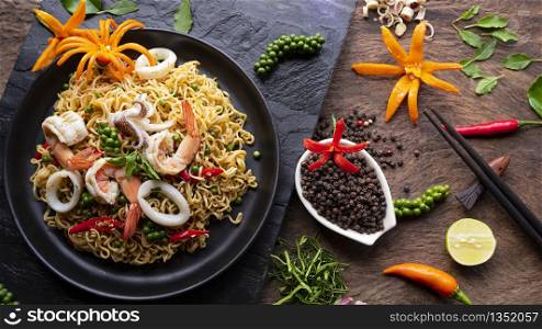 Top view of instant noodles with stir fried spicy seafood and various spices on black stone plate with wooden tabletop, junk food and Asian meal concept