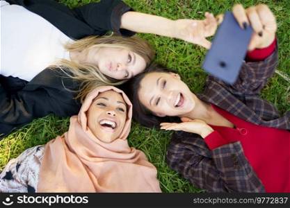 Top view of laughing Muslim female lying near girlfriends pouting lips and taking self portrait on cellphone on grassy ground. Multiracial girlfriends laughing while lying on lawn and taking selfie