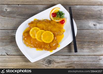 Top view of lemon chicken with tangy sauce and slice lemons on top in white plate with rustic wood underneath.
