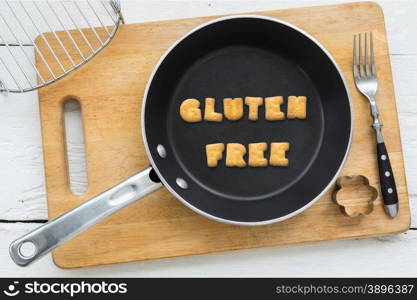 Top view of letter collage made of biscuits. Word GLUTEN FREE putting in black frying pan. Other cooking equipments: fork, cookie cutter and chopping board putting on white wooden table, vintage style image.