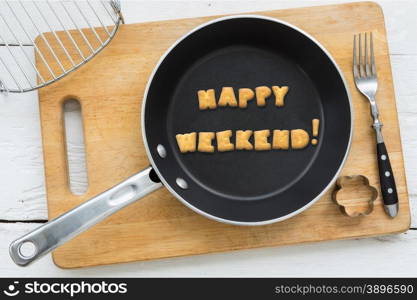 Top view of letter collage made of biscuits. Word HAPPY WEEKEND putting in black frying pan. Other cooking equipments: fork, cookie cutter and chopping board putting on white wooden table, vintage style image.