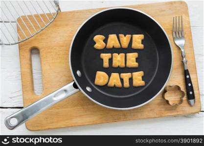 Top view of letter collage made of biscuits. Quote SAVE THE DATE putting in black frying pan. Other cooking equipments: fork, cookie cutter and chopping board putting on white wooden table, vintage style image.