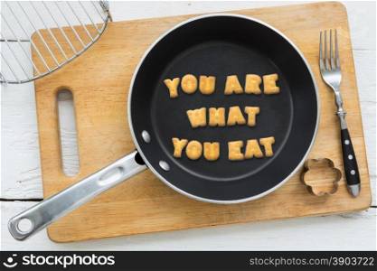 Top view of letter collage made of biscuits. Quote YOU ARE WHAT YOU EAT putting in black frying pan. Other cooking equipments: fork, cookie cutter and chopping board putting on white wooden table, vintage style image.