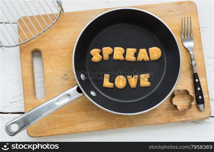 Top view of letter collage made of biscuits. Word SPREAD LOVE putting in black frying pan. Other cooking equipments: fork, cookie cutter and chopping board putting on white wooden table, vintage style image.