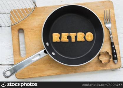 Top view of letter collage made of biscuits. Word RETRO putting in black frying pan. Other cooking equipments: fork, cookie cutter and chopping board putting on white wooden table, vintage style image.