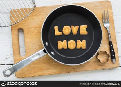 Top view of letter collage made of cookies. Word LOVE MOM putting in black pan. Other kitchen utensils: fork, cookie cutter and cutting board putting on white wooden table, vintage style image.