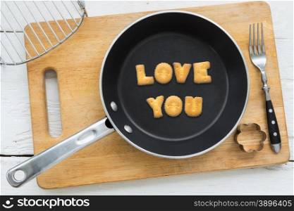 Top view of letter collage made of cookies. Word LOVE YOU putting in black pan. Other kitchen utensils: fork, cookie cutter and cutting board putting on white wooden table, vintage style image.