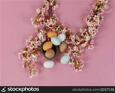 Top view of light artificial cherry blossom flowers with colorful eggs in nest on a soft pink background for a happy Easter holiday concept