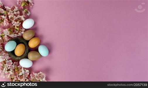 Top view of light artificial cherry blossom flowers with colorful eggs in nest forming left border on a soft pink background for a happy Easter holiday concept