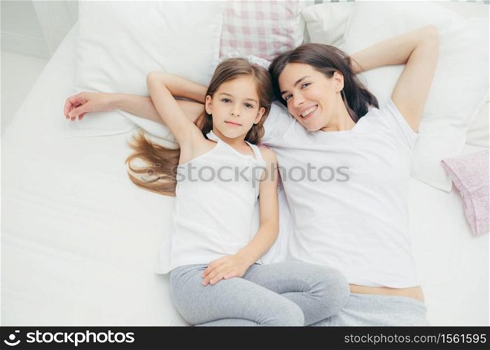 Top view of loving young mother with pleasant smile lies near her small daughter, going to have sleep, enjoy calm domestic atmosphere in bedroom, look directly at camera with joyful expressions