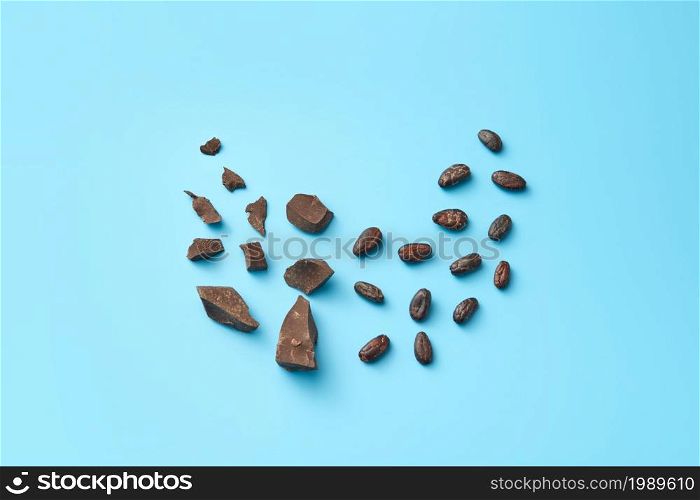 Top view of minimalist composition with pile of organic raw chocolate chunks with cocoa beans on blue background. Chocolate pieces and cocoa beans