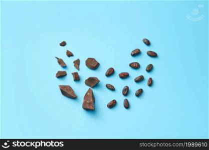 Top view of minimalist composition with pile of organic raw chocolate chunks with cocoa beans on blue background. Chocolate pieces and cocoa beans